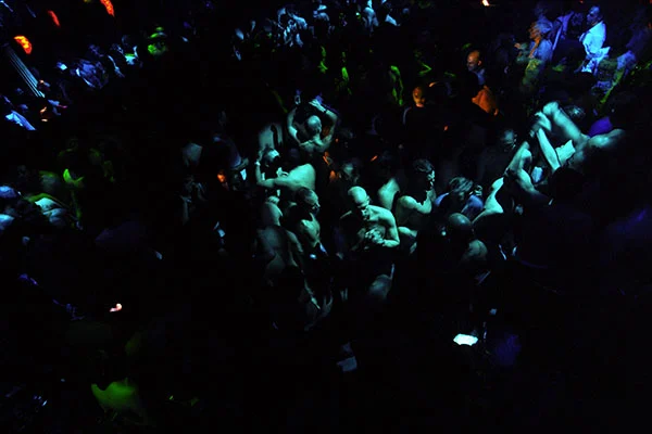 Figures gather in a dark club, interrupted by patches of blue and green light.